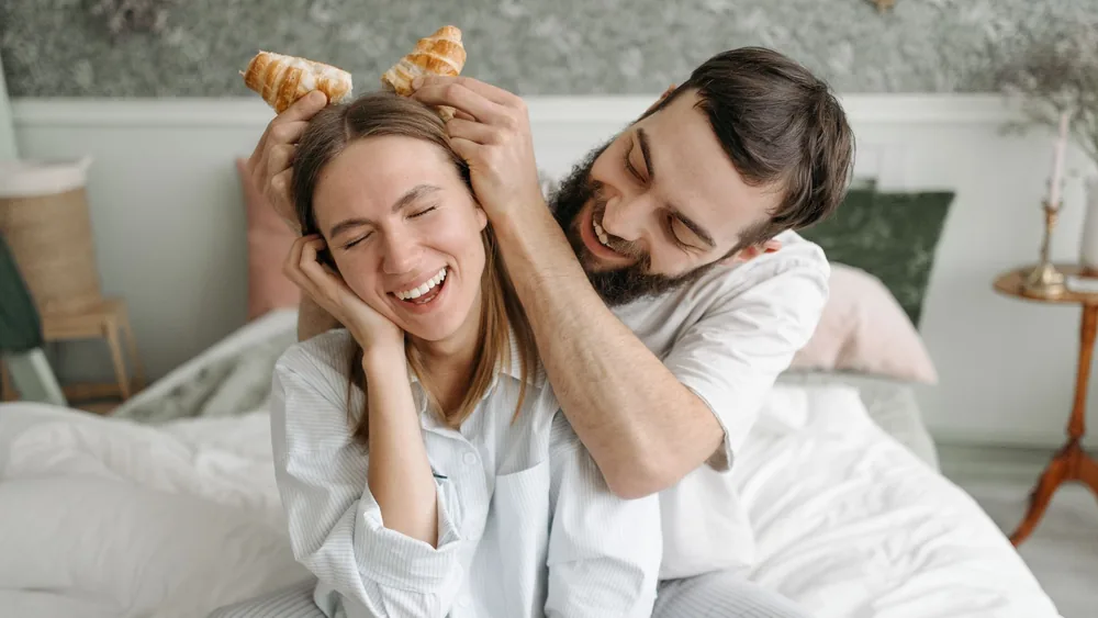 Joyful couple having fun with croissants on a cozy morning in bed.