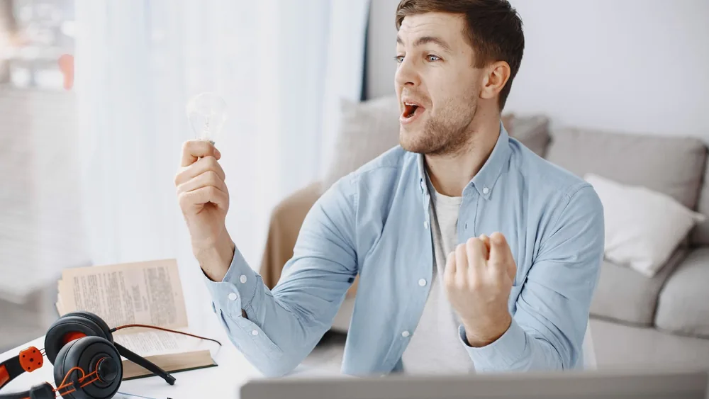 Man joyfully holding a light bulb, symbolizing creativity and innovation in a home office setting.