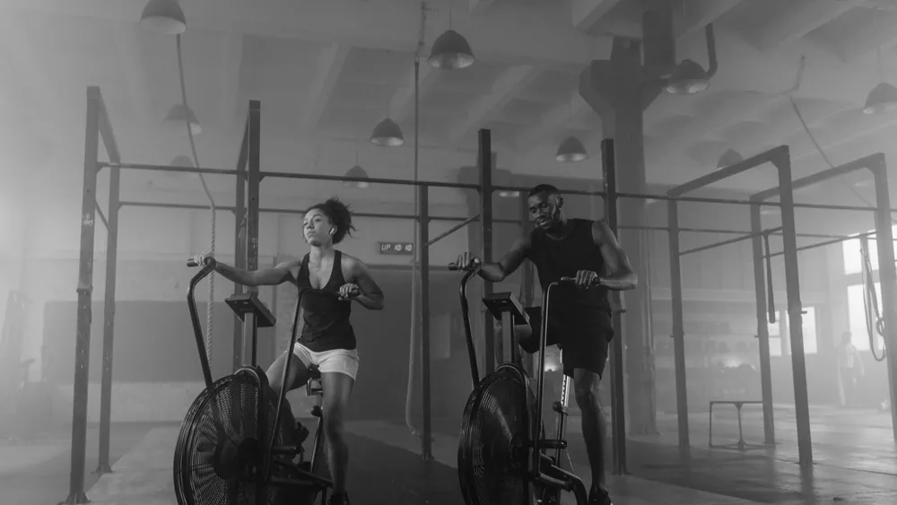 A fit couple exercising on air bikes in a gym setting.