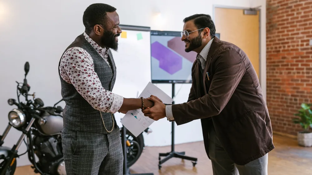 Two male professionals shaking hands in a modern office setting, symbolizing teamwork and collaboration.