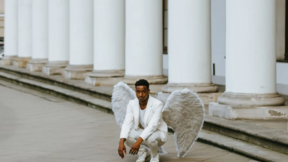 A man in a white suit with angel wings kneels elegantly outdoors against a column backdrop.