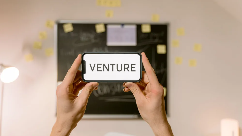 A smartphone displaying 'Venture' held by hands in an office setup. Ideal for business and startup themes.