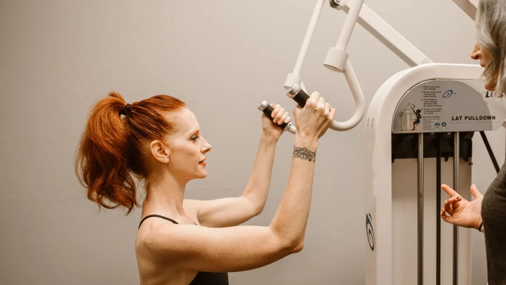 Woman working out on lat pulldown machine while being guided by a trainer.