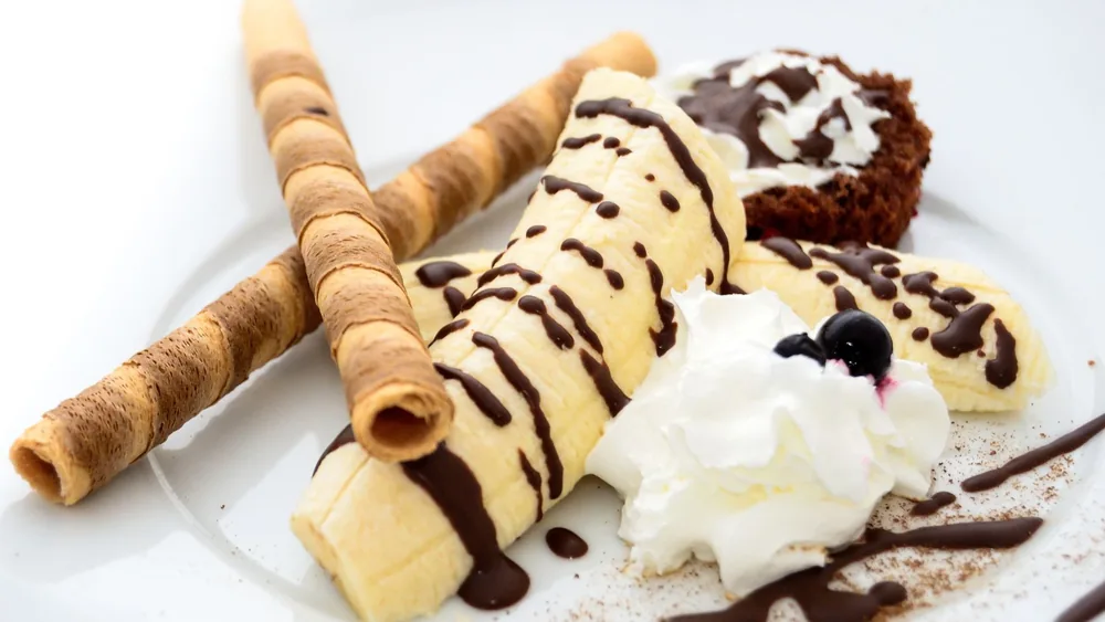 Close-up of a banana dessert with chocolate drizzle, whipped cream, and wafer sticks.