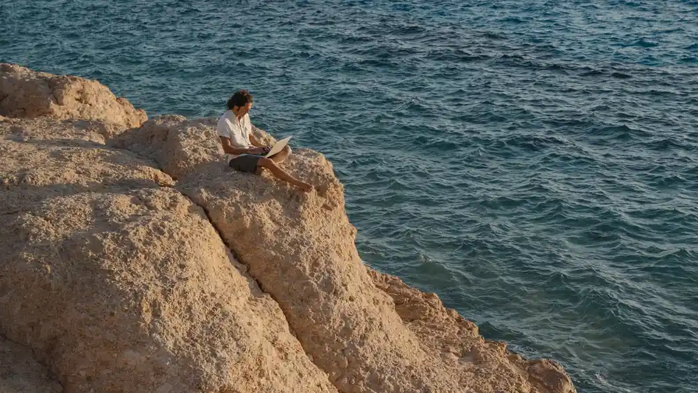 A man sits on a rocky cliff by the sea, working remotely on a laptop.