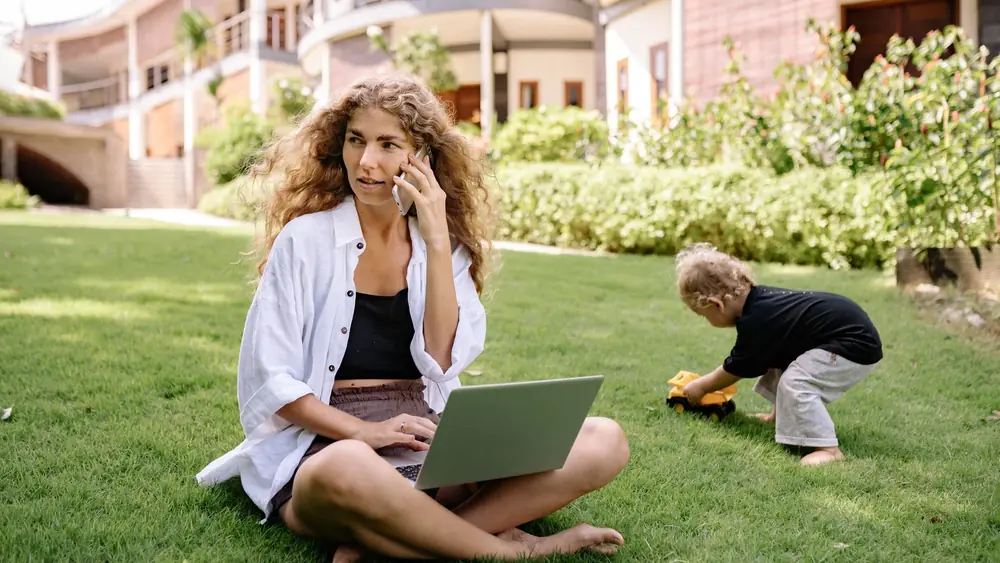 Woman multitasking on phone and laptop while child plays outdoors.