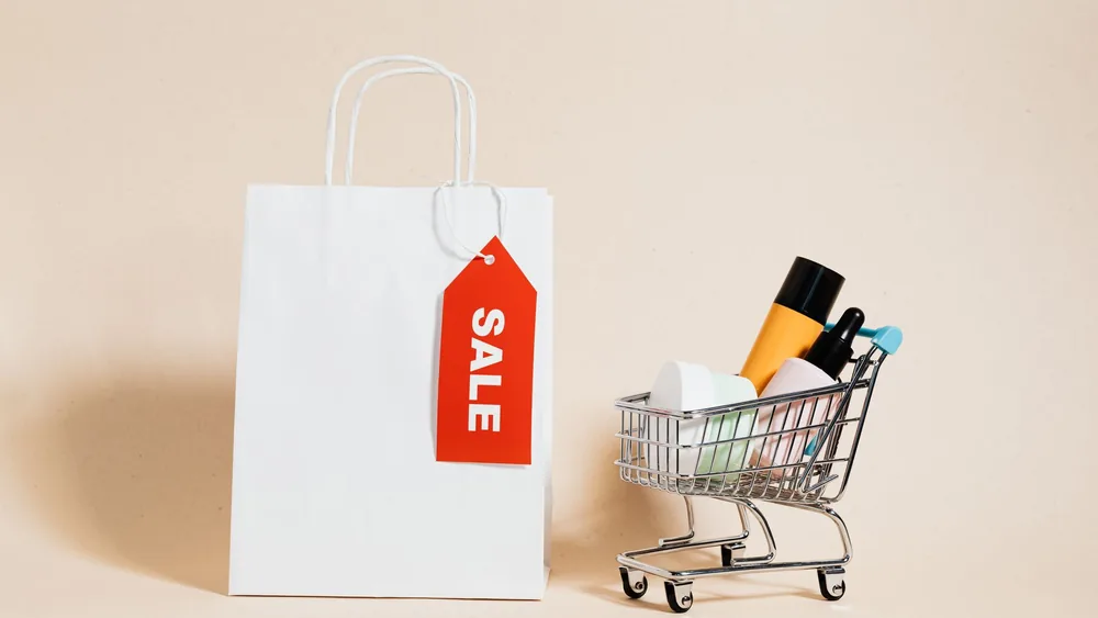 White shopping bag with red sale tag and mini shopping cart on beige background.