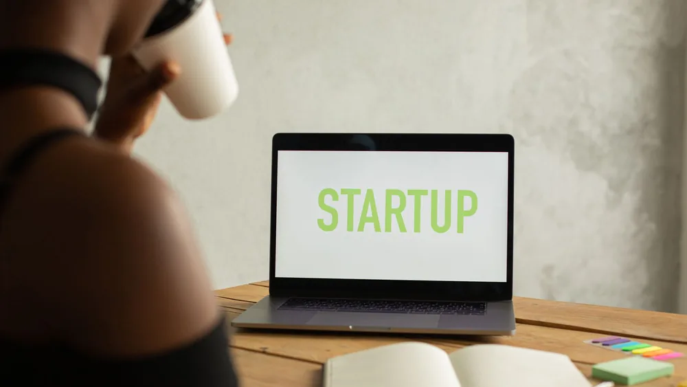 Crop anonymous black female entrepreneur sitting at table with opened netbook with startup on screen and notebook and drinking coffee from takeaway cup while working on project remotely