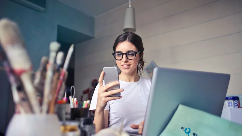 A woman using her phone at a desk, surrounded by art supplies and a laptop, in a creative workspace.