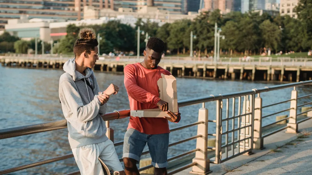 Positive diverse male friends wearing casual clothes sharing delicious pizza while standing near embankment handrail in urban city