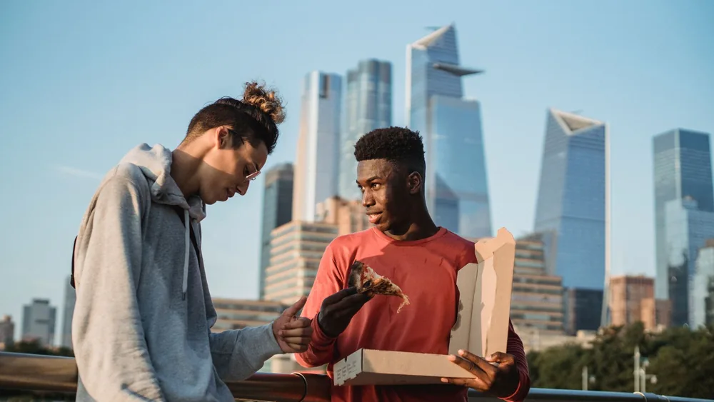 Content black man offering yummy pizza to male friend while spending summer day on modern city embankment