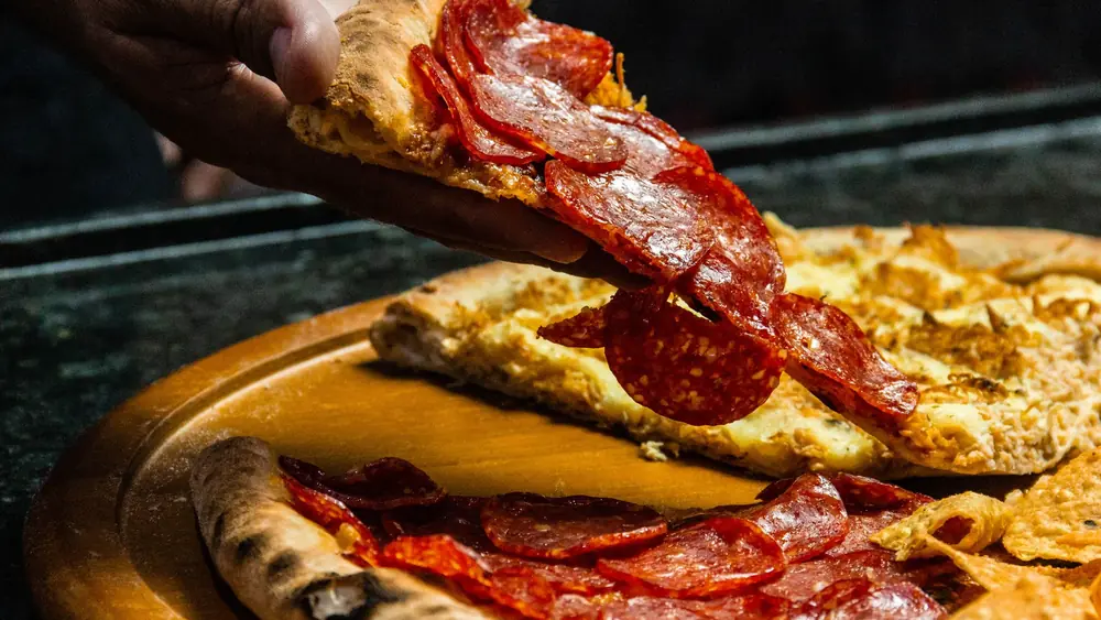 Fresh pepperoni pizza slice being lifted from a wooden board by hand. Tasty and mouth-watering dinner option.