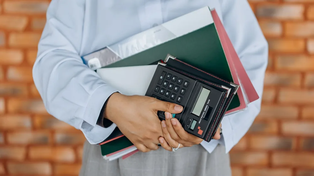Close-up of a person holding a calculator and folders against a brick wall background.