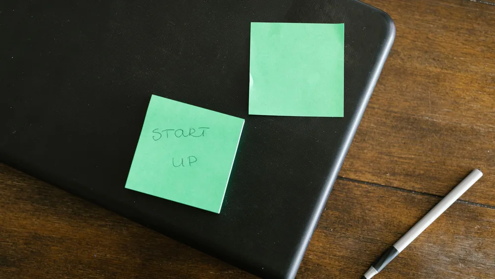 A laptop with green sticky notes and a pen on a wooden desk, symbolizing startup planning.