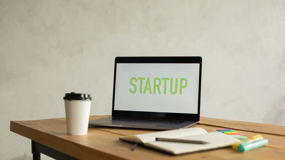 Opened netbook with Startup title on screen and white takeaway cup of coffee placed on wooden table near opened notebook against white wall