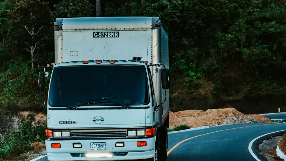 A white truck driving on a winding road surrounded by dense green forest plants.
