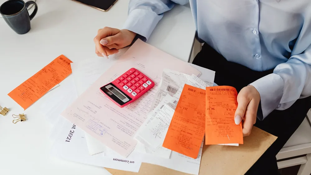 A woman reviews receipts and calculates expenses at a desk with a pink calculator.