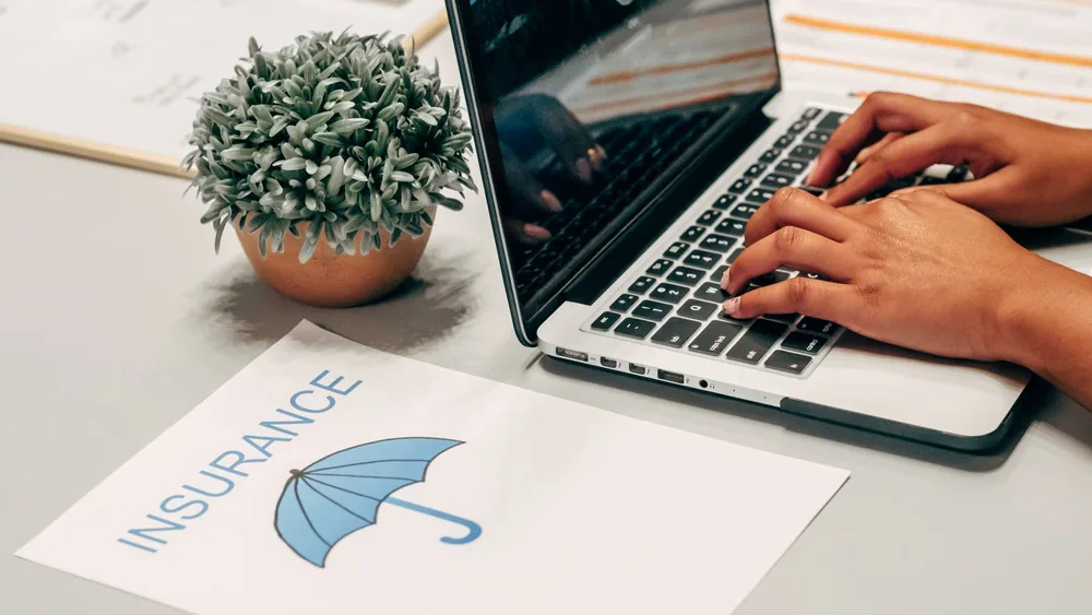 Hands typing on a laptop at a desk with an insurance paper and plant, suggesting a work environment.