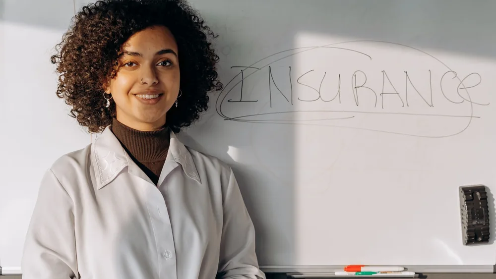 African American woman smiling in office setting with a whiteboard displaying 'Insurance'.
