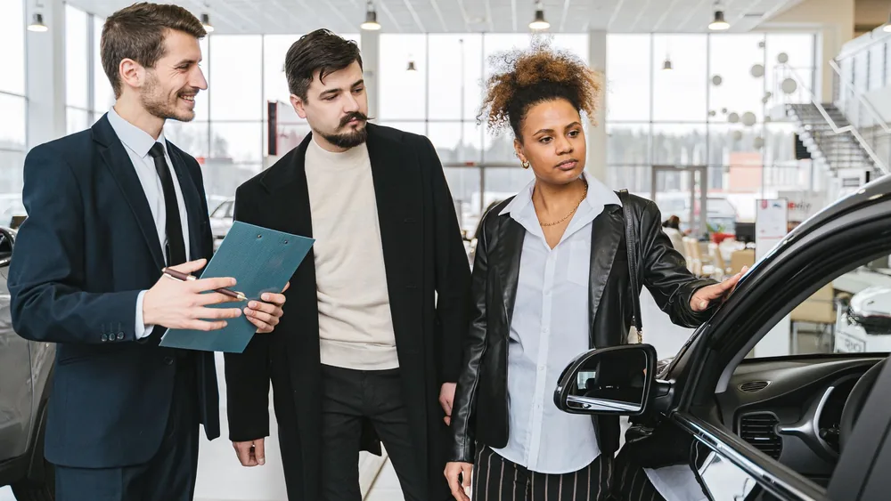 An interracial couple consults with a salesman at a car dealership, exploring vehicle options.