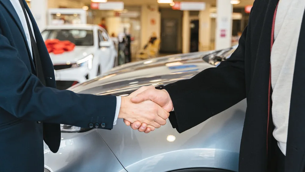 Two businessmen shaking hands in a car dealership, sealing a deal.