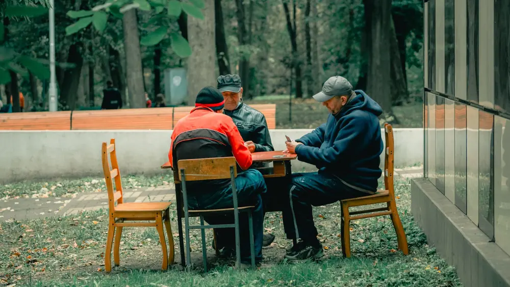 Three men sitting around a table playing cards in a park setting surrounded by trees.