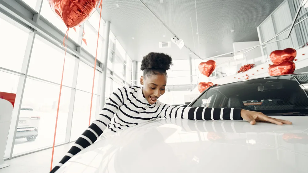 Excited woman embraces her new car at a dealership, surrounded by red balloons.
