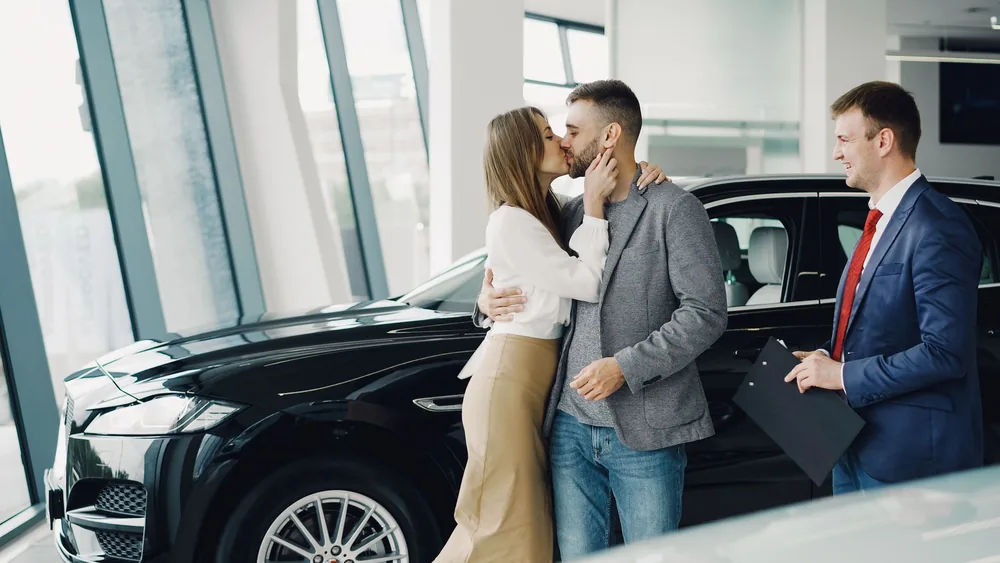 Excited young couple kissing after purchasing a new car at a modern showroom.