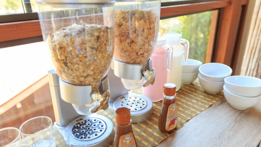 Close-up of cereal dispensers and breakfast setup with bowls and milk on a wooden table.