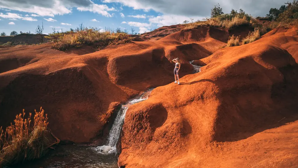 Explore the vibrant red sand landscapes and waterfall of Kauai, Hawaii on a bright day.