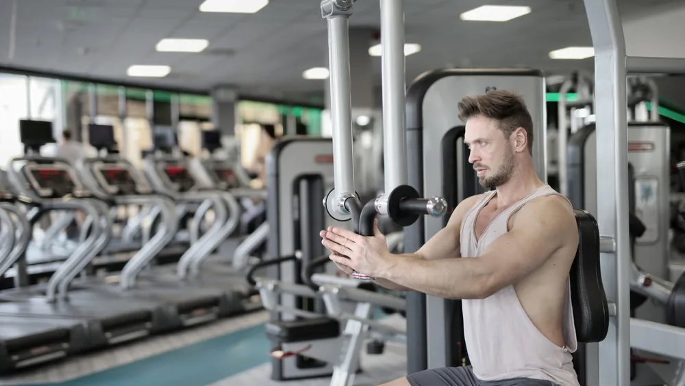Serious strong male athlete in casual sportswear sitting on pec deck machine and performing exercise bringing hands together and looking forward in modern fitness center