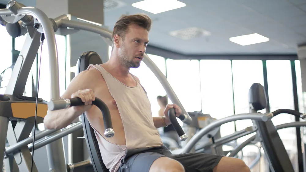 Focused adult male athlete in sportswear using weight machine for arms and chest during workout in modern spacious fitness center in morning