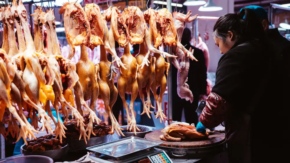 A market vendor prepares poultry at a traditional indoor Asian marketplace with hanging chickens.