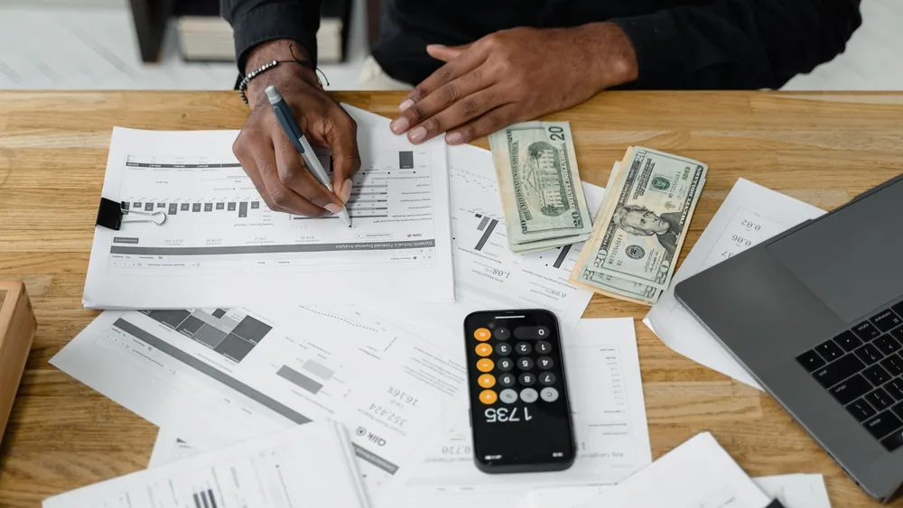 Man working on financial reports with calculator, money, and laptop on a desk.