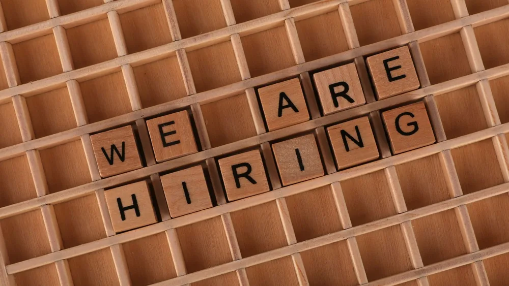 Wooden letter tiles arranged to spell 'We Are Hiring' on a wooden grid background.