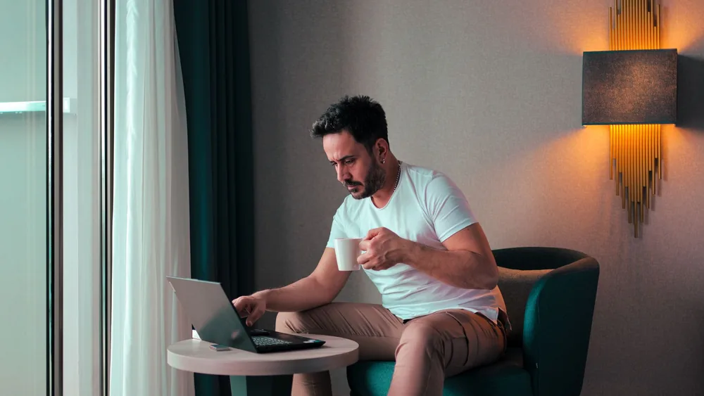 A man sits in a cozy hotel room in Cyprus, working on a laptop while enjoying a coffee.
