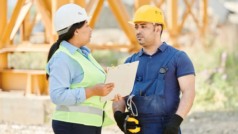 Male and female construction workers talking with safety gear at an outdoor site.