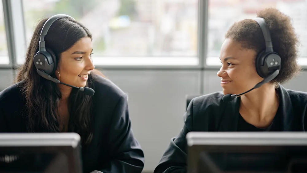 Two smiling female call center agents with headsets engaging in teamwork.