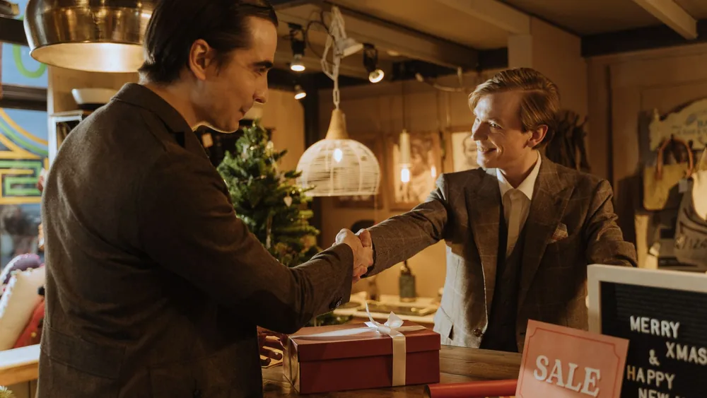 Two men in suits shake hands over a holiday gift in a cozy store ambiance.