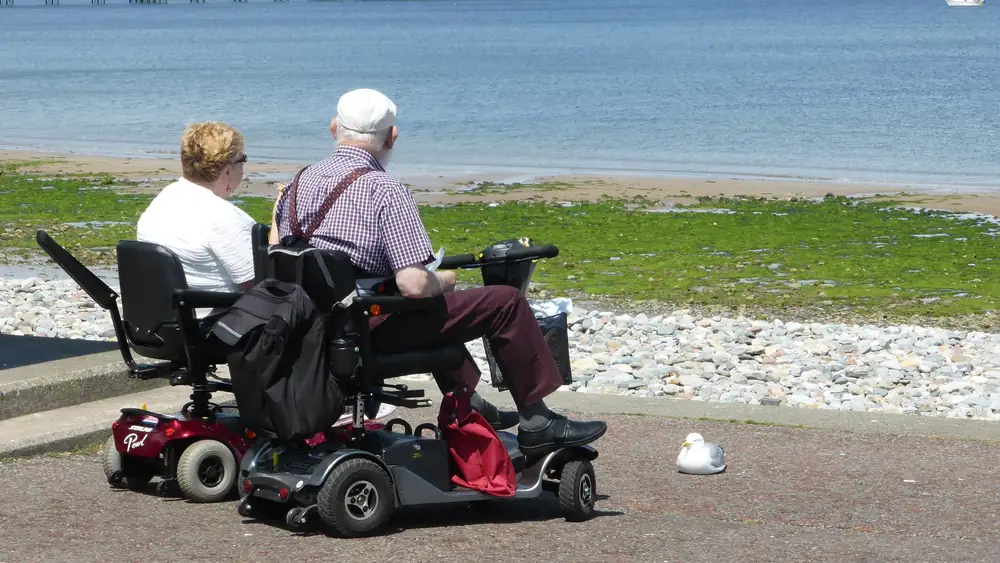 An elderly couple enjoys a seaside view while seated on mobility scooters, emphasizing leisure and accessibility.