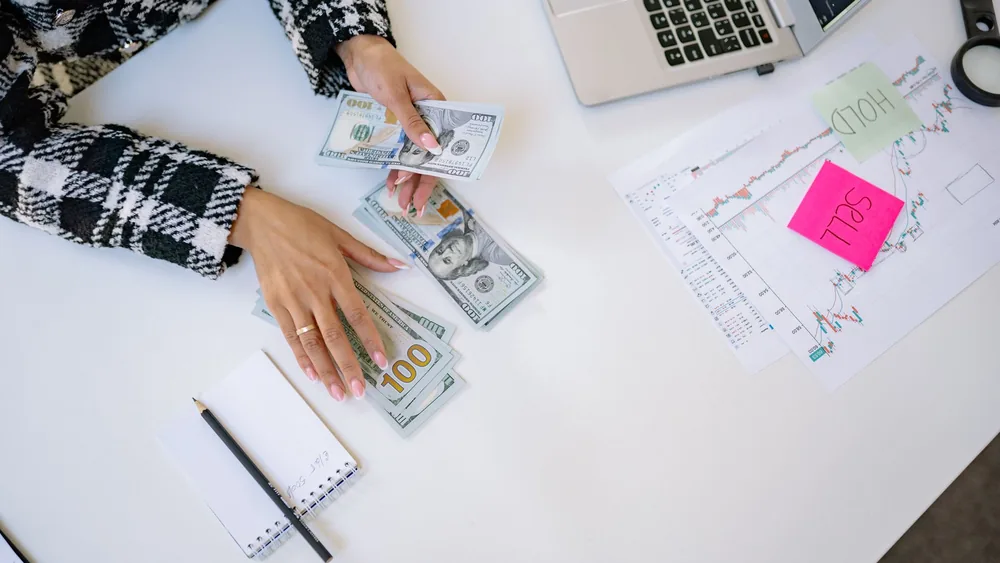 Businesswoman counting dollar bills with financial charts and laptop on table. Investment and finance concept.