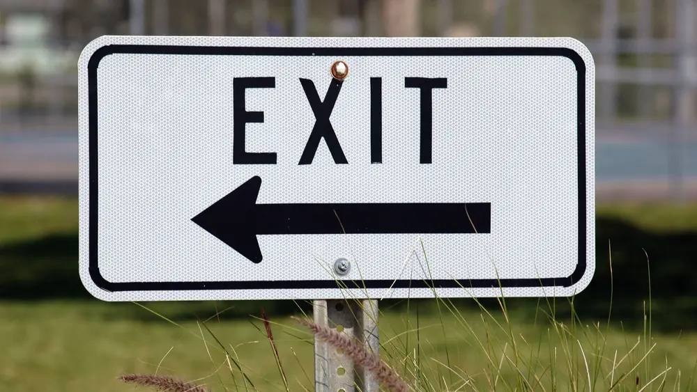 Exit sign with left-pointing arrow in a grassy outdoor setting.