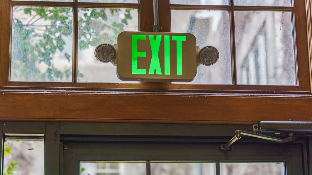 Green exit sign illuminated above a glass doorway indoors with natural light.