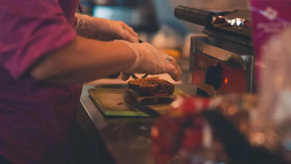 A chef wearing gloves prepares a sandwich indoors with professional equipment.