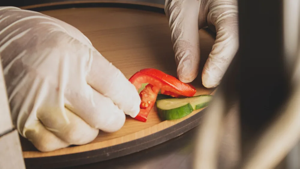 Close-up of gloved hands arranging cucumber and tomato slices on a wooden tray.