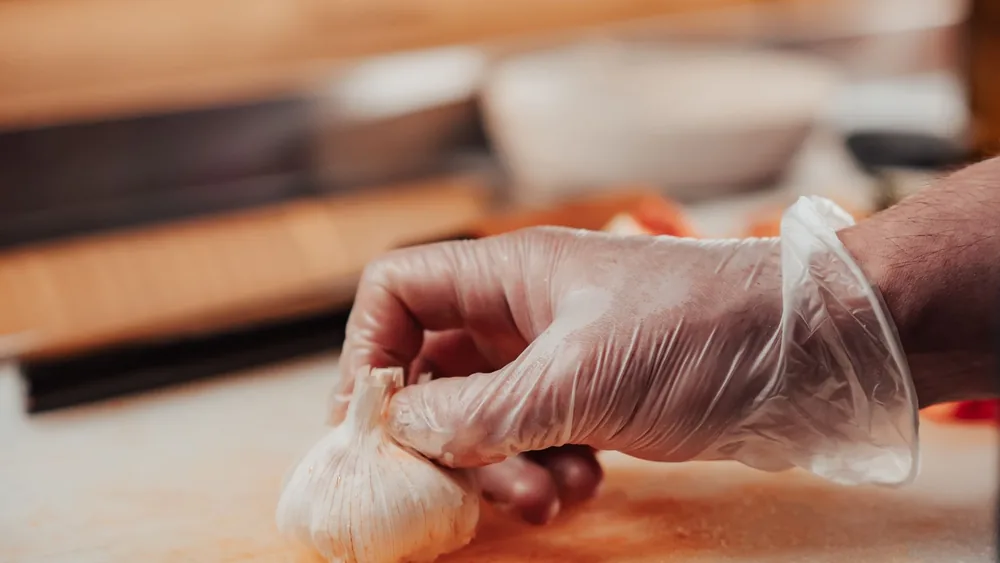 Chef's hand in glove holding garlic bulb in a kitchen setting, ready for cooking.