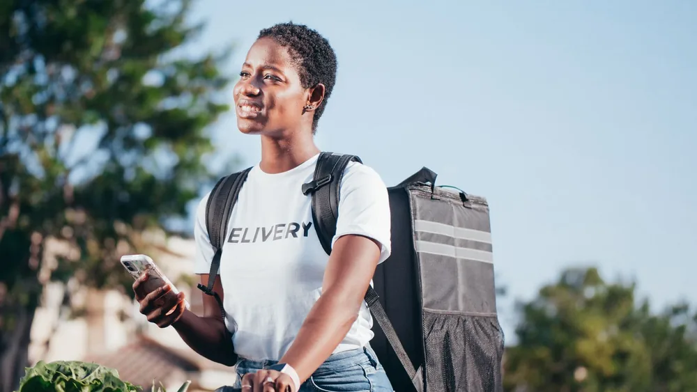 Happy deliverywoman in uniform holding phone and backpack in sunny outdoor setting.