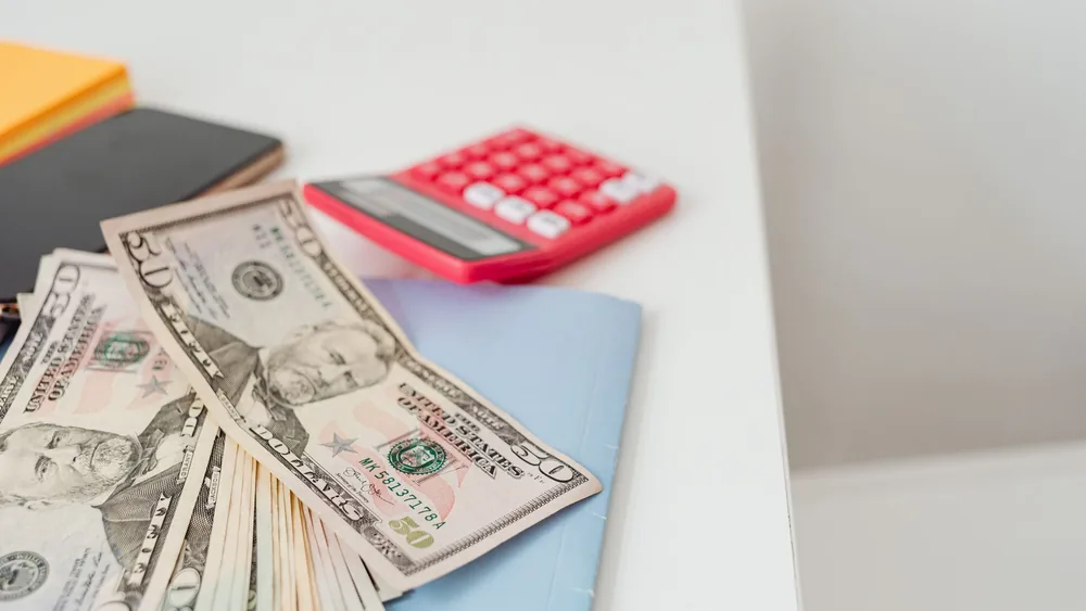 Close-up of notebooks, US dollars, and a calculator on a desk.