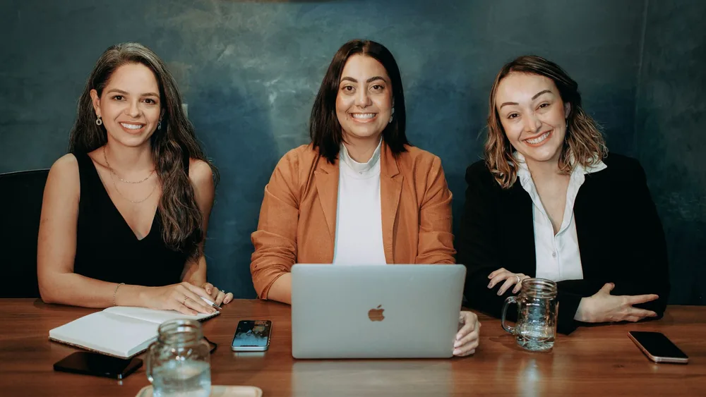 Three professional women sitting at a wooden desk in an office with laptops and notebooks.