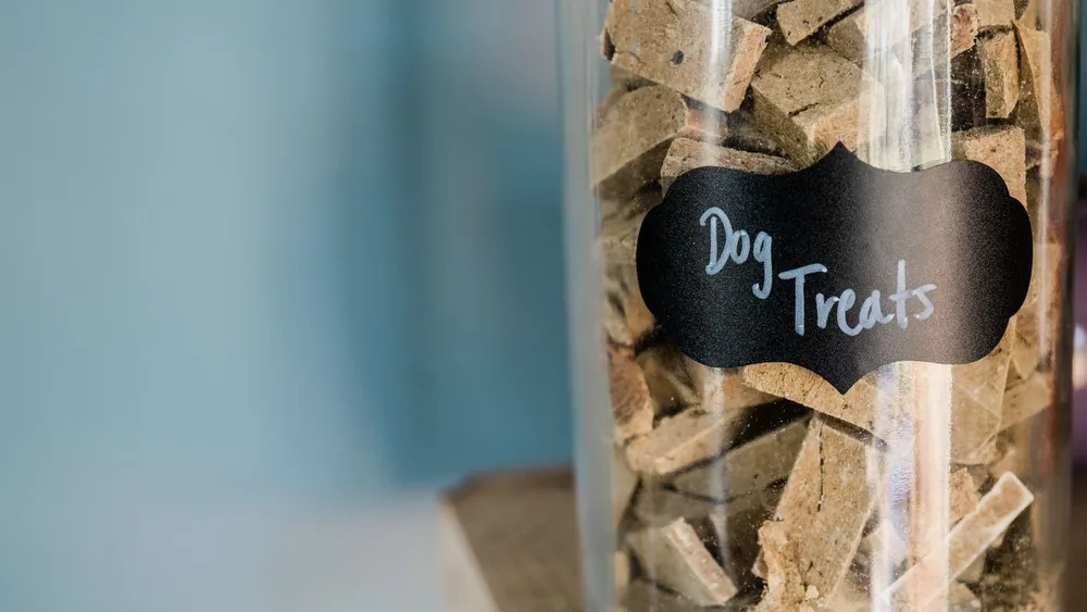 A close-up image of dry dog treats stored in a labeled glass jar on a wooden surface.